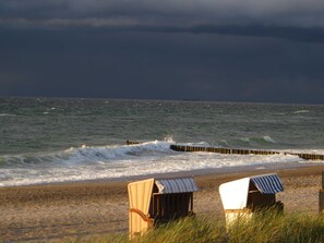 Two beach chairs with striped awnings on a sandy beach, facing the ocean with waves crashing onto a wooden breakwater.