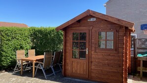 A wooden cabin with a door and windows, a table with chairs, and a brick wall in the background.