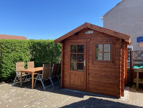 A wooden cabin with a door and windows, a table with chairs, and a brick wall in the background.