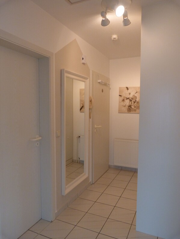A hallway with white walls, a tiled floor, and a door leading to a bathroom. There is a ceiling light fixture and a radiator on the wall. - Comfortable apartment with 2 bedrooms, balcony, WiFi, renovated in 2016 (Grömitz)