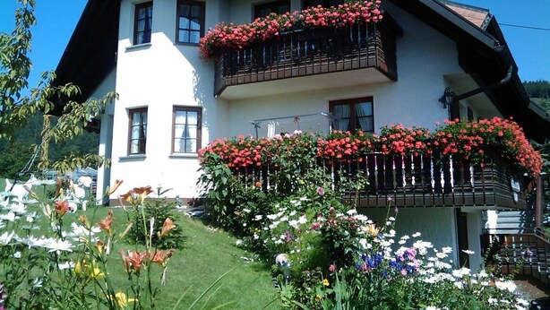 A two-story house with a white facade and dark brown roof. It features a balcony with red flowers and a well-maintained garden with various plants and flowers. - Beautiful, large, comfortable vacation apartment to feel at home in. (Dachsberg (Südschwarzwald))