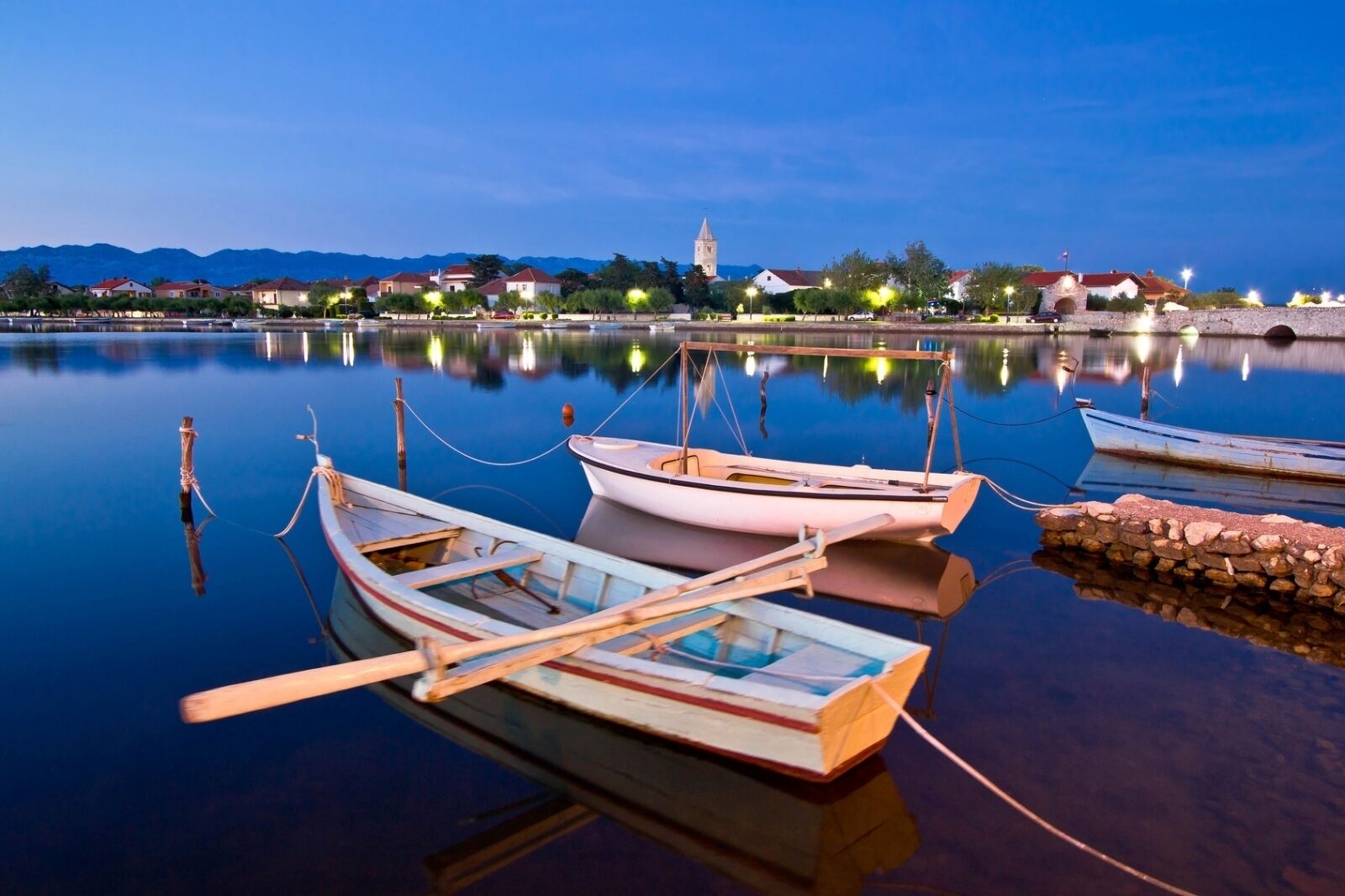 Um lago tranquilo com barcos atracados, uma torre de igreja ao fundo e casas ao longo da margem.