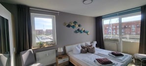 Ein Hotelzimmer mit einem Bett, einem Nachttisch, einem Sessel, einem Fenster mit Blick auf Gebäude und einer Wanddekoration.