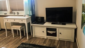 Una habitación con un escritorio blanco, televisión, radiador y ventana con persianas.