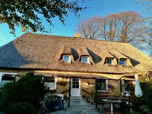 Un bâtiment à toit de chaume, doté d’un porche, de plantes en pot et de sièges d’extérieur.
