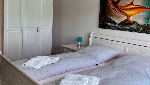 Un dormitorio con cama, armario, mesita de noche, lámpara y un cuadro en la pared.