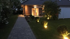 Et hus med en belagt indkørsel, der fører til en hoveddør, en lille have med buske og en bænk, samt en garage.