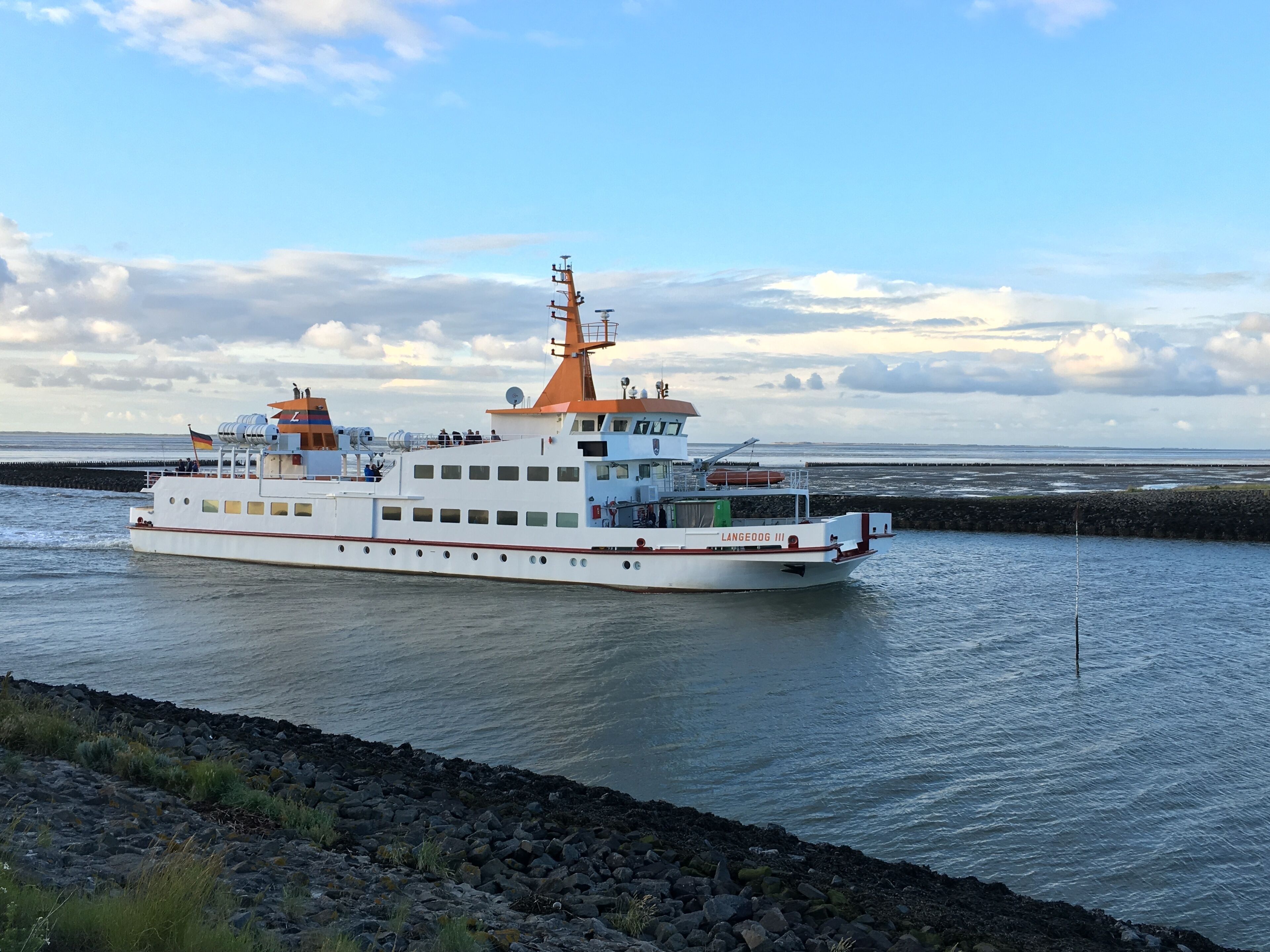 Un ferry con varias cubiertas y una chimenea anaranjada destacada, navegando por una masa de agua con una costa rocosa en primer plano.