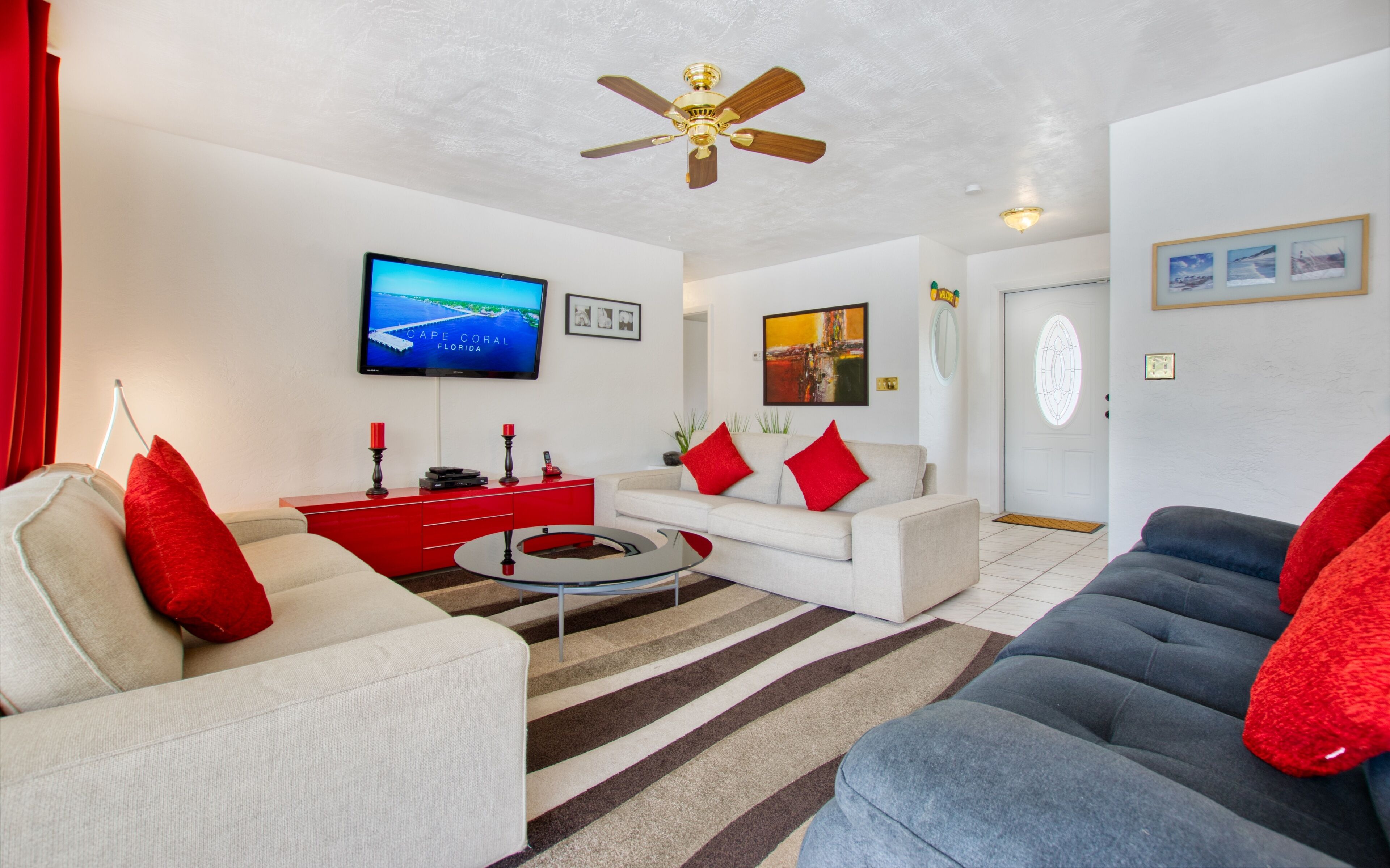 Sala de estar com televisão exibindo uma paisagem, ventilador de teto e sofá vermelho e branco.