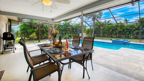 A covered outdoor dining area with a table set for two, chairs, a ceiling fan, and a view of a swimming pool and tropical vegetation. - Holiday home with large heated pool - south exposure (Cape Coral)