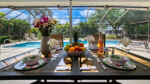 An outdoor dining area with a pool view, featuring a table set with a fruit bowl, a bottle of wine, and a vase of flowers. - Holiday home with large heated pool - south exposure (Cape Coral)