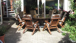 A round glass table with wooden chairs under a canopy, surrounded by potted plants and a brick patio.
