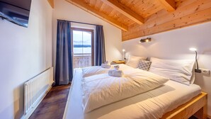 Ein Schlafzimmer mit Holzbalkendecke, einem großen Bett mit weißer Bettwäsche, einem Nachttisch, einer Lampe und Blick durch ein Fenster mit Vorhängen.