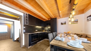 Eine moderne Küche mit dunklen Schränken und einer Holzbalkendecke, ein Essbereich mit einem Holztisch für vier Personen und Blick auf den angrenzenden Wohnraum.