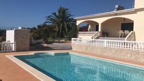 A swimming pool with a stone wall and white balustrade, a staircase leading to a covered patio with outdoor furniture, and a palm tree in the background. - Detached villa with sea view, heated pool and air conditioning (Caramujeira)