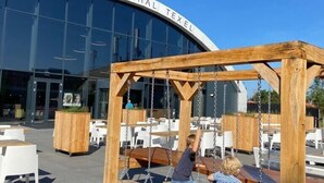 Un espace extérieur aménagé avec des balançoires en bois et des tables, situé devant un bâtiment à la façade vitrée, sur lequel on peut lire l'inscription « EVENEMENTENHAL TEXEL ».