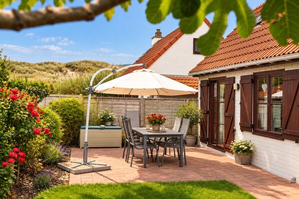 A patio with a table and chairs, a white umbrella, and a garden with red flowers.