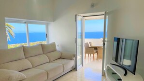 Una sala de estar moderna con un sofá color beige, un televisor de pantalla plana y un ventanal de vidrio que da a un balcón con vistas al mar.