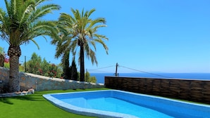 Una piscina con muro de piedra y palmeras, rodeada por un muro de piedra y una valla de madera, con vista al mar.
