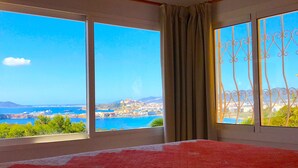 Un dormitorio con una cama grande, dos ventanas con cortinas y vistas a una ciudad costera.