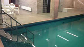 En indendørs swimmingpool med flisegulv, en trappe ned til poolen og et opholdsområde med stole og et bord.