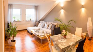 A modern living room with a sofa, coffee table, and dining table, featuring a view of the outdoors through large windows. - Relaxation in the vineyards - Ferienwohnung Isinger (Rüdesheim am Rhein)