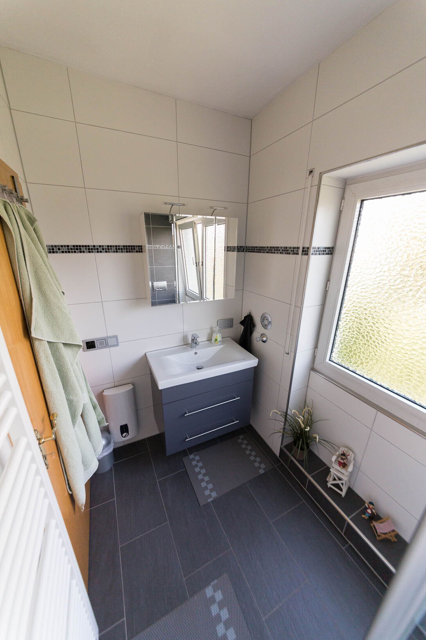Un baño con un lavabo blanco, baldosas oscuras en el suelo, un espejo y una ventana.