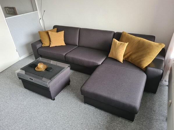 Un sofá modular con dos cojines color mostaza, un pequeño reposapiés y una mesa de centro de vidrio con un adorno.