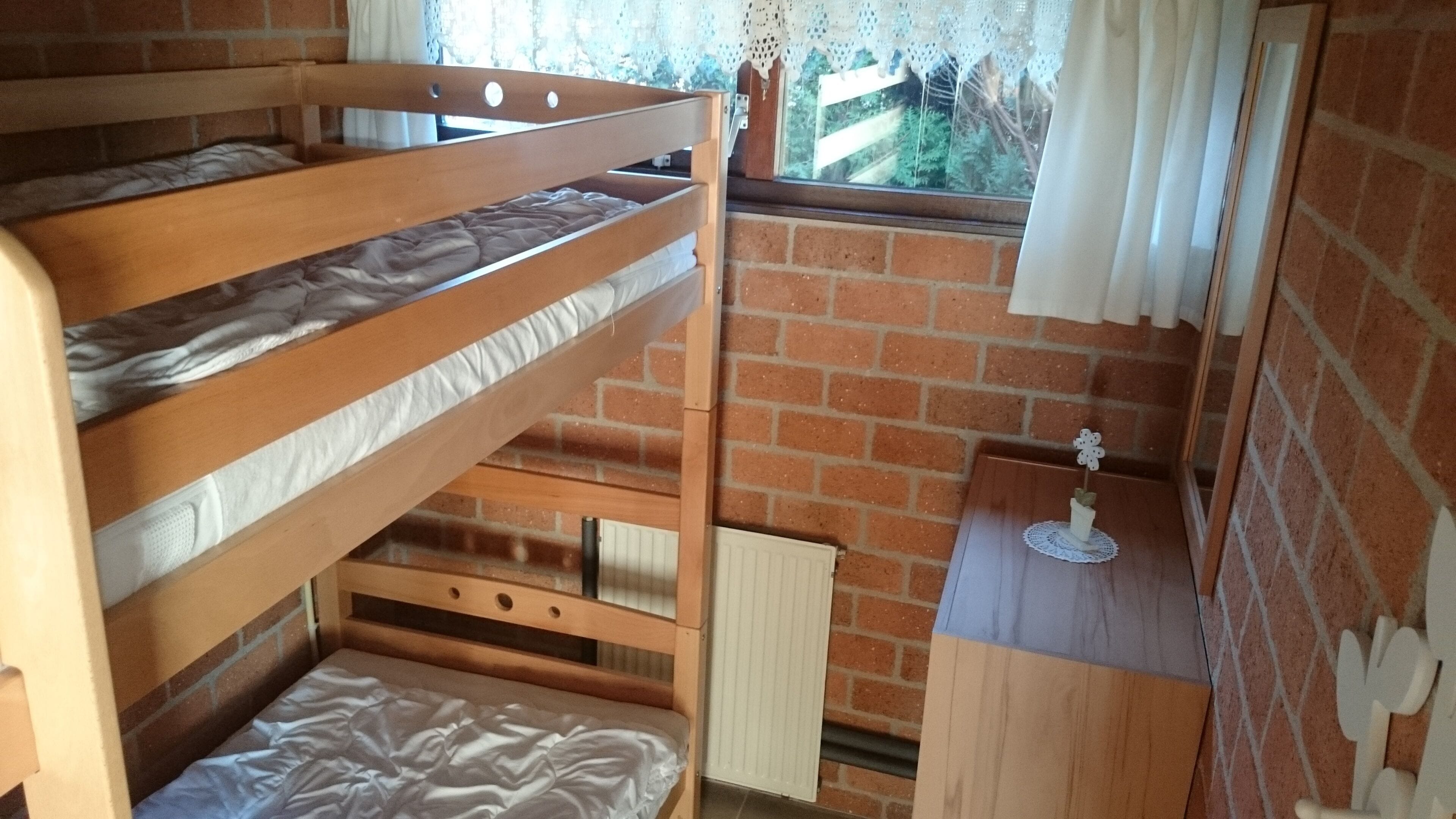 Une chambre avec un lit superposé, un mur en briques, une fenêtre ornée de rideaux en dentelle et une petite table sur laquelle est posé un objet décoratif.