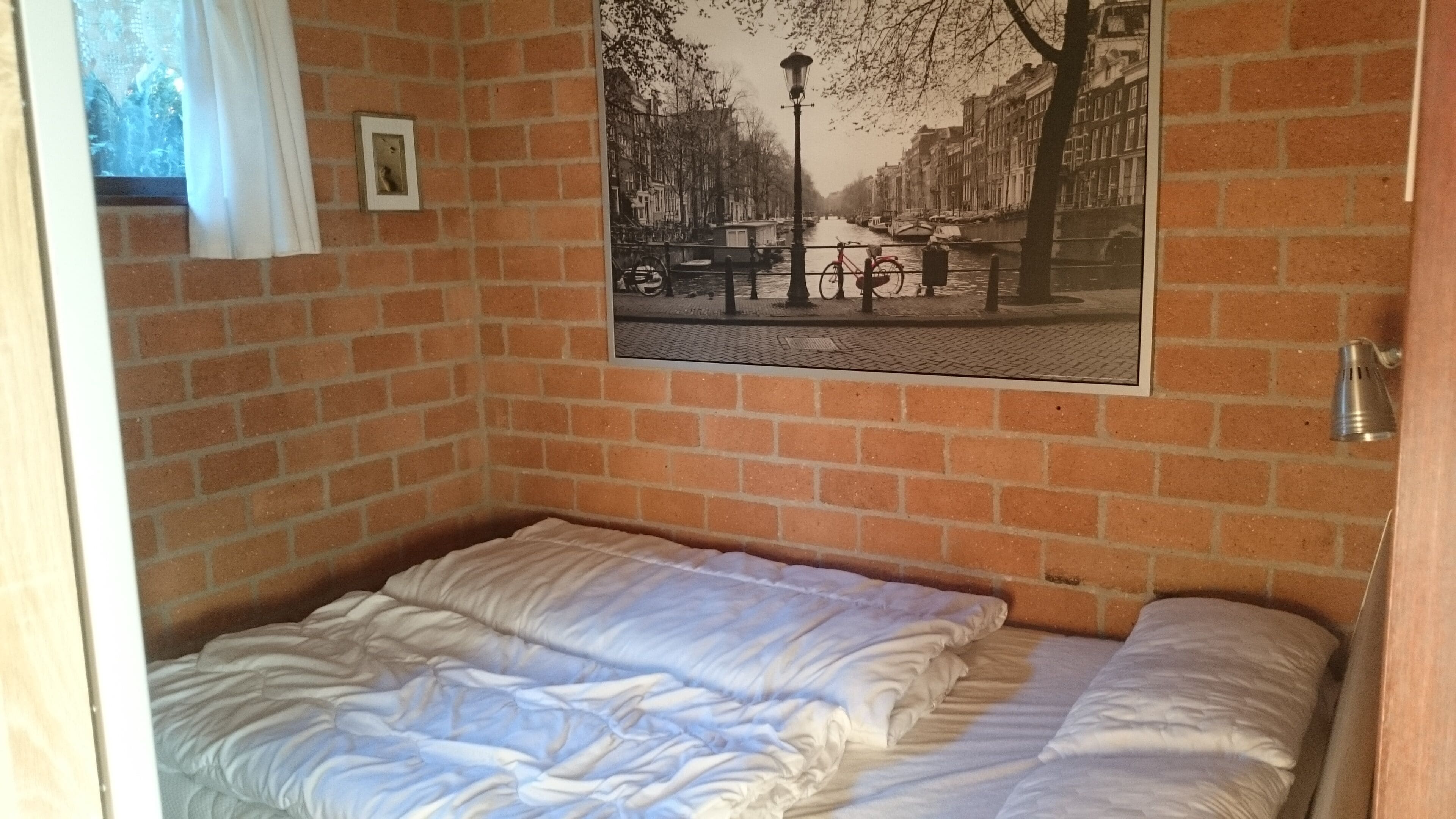 A small, single-bed room with a brick wall and a framed picture of a canal scene.