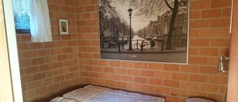 A small, single-bed room with a brick wall and a framed picture of a canal scene.