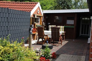 Eine Terrasse mit Holztisch und -stühlen, ein kleiner Garten mit verschiedenen Pflanzen und Blumen sowie eine Ziegelmauer.
