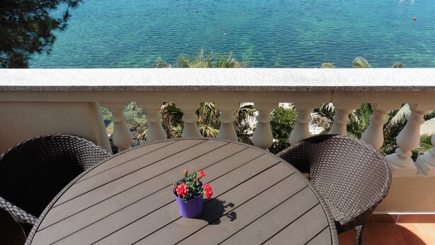 Un balcón con una mesa redonda y sillas de mimbre, con vista a un mar azul despejado y barcos.