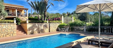 A swimming pool with lounge chairs and an umbrella, surrounded by stone walls and palm trees.