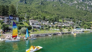 A lakeside view with a sailboat, a speedboat, and a dock with boats. There are buildings in the background and a mountainous landscape.