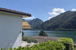 A terrace with a view of a lake and mountains, a building with a number sign, and outdoor furniture.