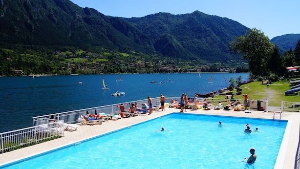 A swimming pool with people swimming and sunbathing, surrounded by mountains and a lake.