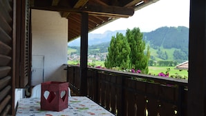 A balcony with a table covered in a floral tablecloth, a wooden railing, and a view of a green landscape with mountains in the background. - Apartment with panoramic views (Halblech)