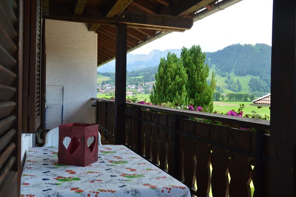 A balcony with a table covered by a floral tablecloth, a red decorative box, and a view of a green landscape with trees and a mountain in the distance. - Apartment with panoramic views (Halblech)