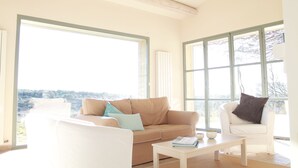 A spacious living room with a beige sofa, a white armchair, and a coffee table. Large windows provide a view of the outdoors.