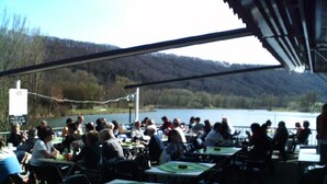 An outdoor dining area with tables and chairs, overlooking a body of water and hills. - Ideal nature: sunny home between a swimming lake, forest & the Altmühl River (Bayern)