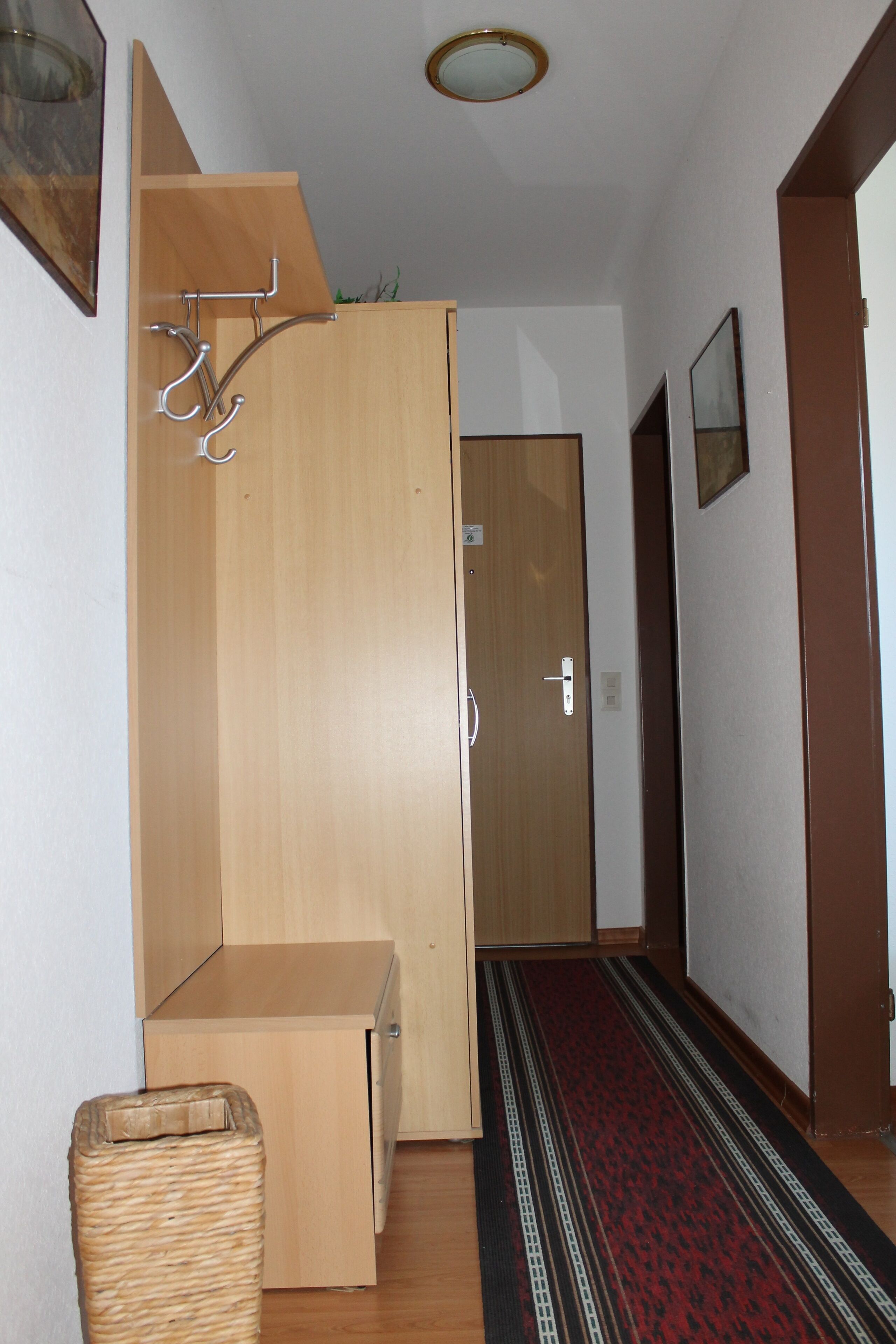Ein Flur mit einem hölzernen Kleiderschrank, einem kleinen Korbwäscheschrank und einem roten Teppich mit Muster.