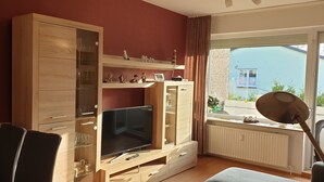 Een moderne woonkamer met een televisie, een houten kast en een raam met uitzicht op een gebouw.