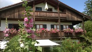 Un chalet traditionnel en bois, doté de balcons ornés de fleurs roses, d’un parasol blanc et surplombant un ciel d’un bleu limpide.