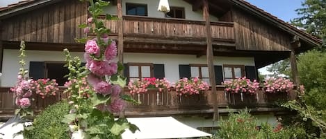 Un chalet traditionnel en bois, doté de balcons ornés de fleurs roses, d’un parasol blanc et surplombant un ciel d’un bleu limpide.