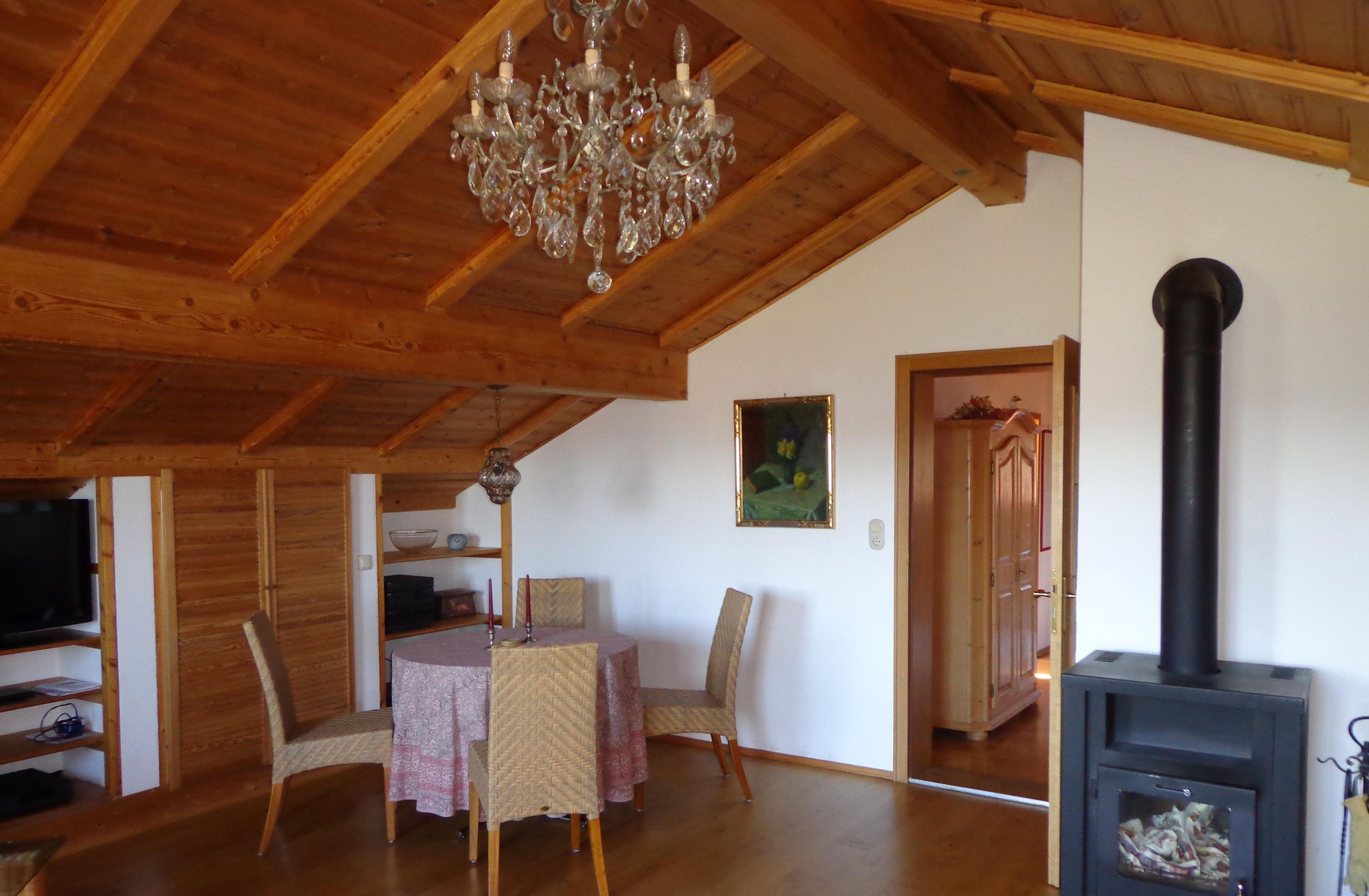 Ein Holzinterieur mit einem Esstisch, Stühlen, einer Chandelüster, einem Gemälde und einem Kaminofen.