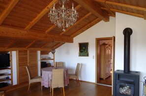 Un intérieur en bois comprenant une table à manger, des chaises, un lustre, un tableau et un poêle à bois.