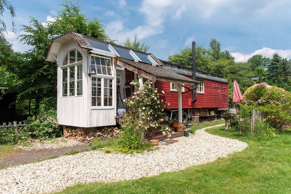 A red wooden house with a white annex, surrounded by a garden with gravel paths and various plants. - Circus wagon by the river (Windeck)
