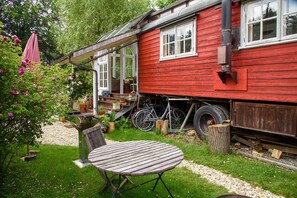 A red wooden house with a porch, a bicycle, and a table in a garden. - Circus wagon by the river (Windeck)
