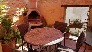 Une terrasse avec une table ronde et des chaises en osier, un four en briques et des plantes en pot.